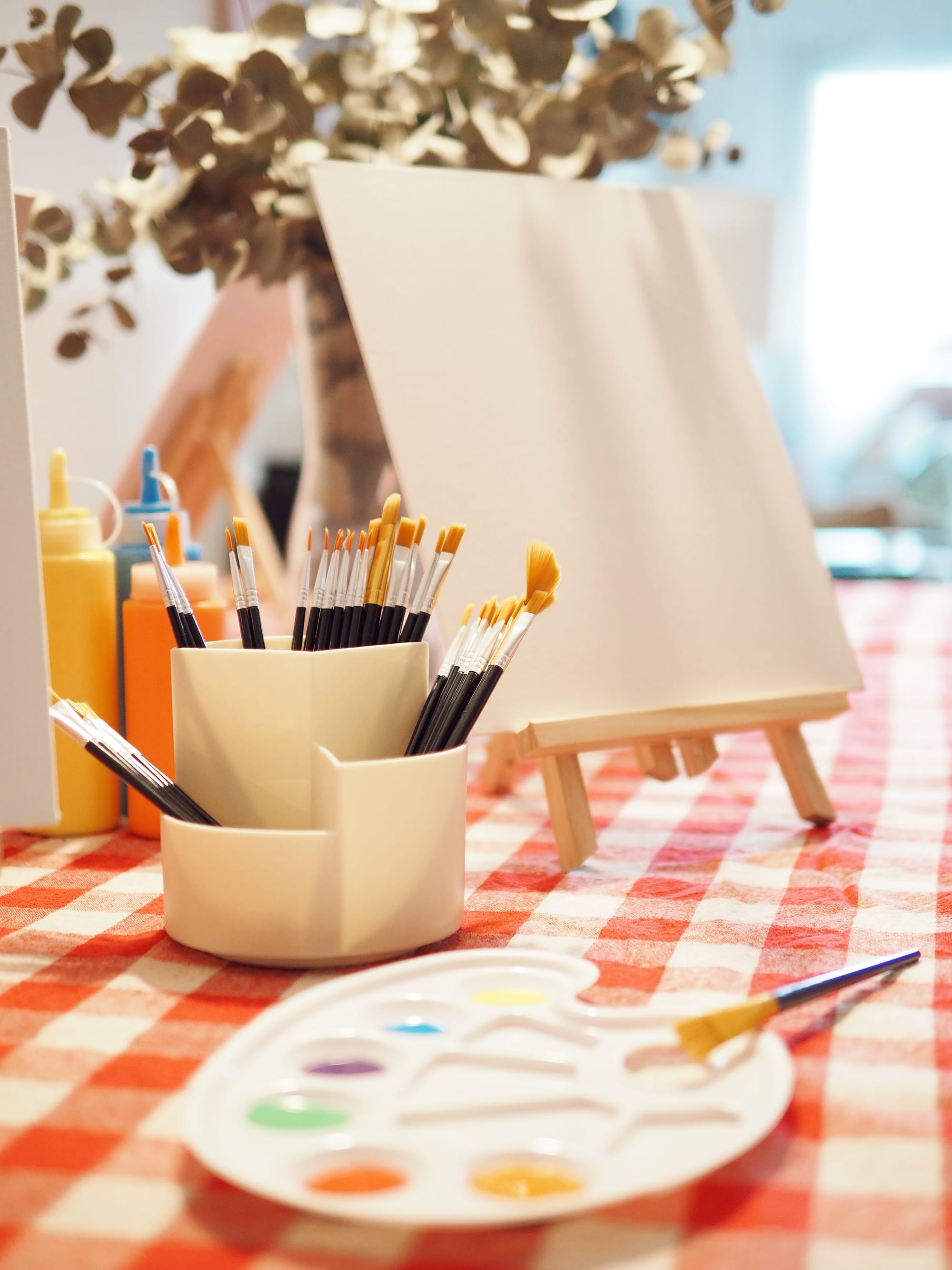 An easel with a canvas, a pot of brushes and a palette on a red gingham cloth